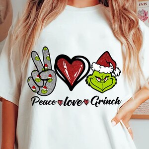 Puede incluir: Camiseta blanca con un diseño festivo. El diseño incluye las palabras "Peace love Grinch", un signo de la paz con caras de Grinch, un corazón rojo brillante y la cara del Grinch con un gorro de Papá Noel.