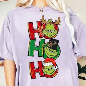 Puede incluir: Una camiseta lila con la palabra "HO" repetida tres veces, cada "O" reemplazada por una cara del Grinch. El Grinch lleva accesorios festivos: cuernos de reno, sombrero de copa y gorro de Papá Noel. Brillo dorado acentúa el diseño.