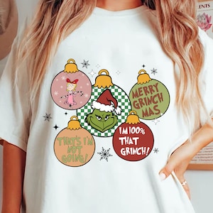 May include: White t-shirt with five Christmas bauble designs featuring Grinch characters and quotes. The baubles display the Grinch, Cindy Lou Who, and phrases such as "Merry Grinch Mas" and "I'm 100% That Grinch!"