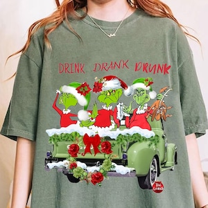 Puede incluir: Camiseta verde con un gráfico de los personajes de Grinch en la caja de una camioneta verde, con el texto "DRINK DRANK DRUNK" en rojo. Los Grinch llevan gorros de Papá Noel y trajes rojos. Una corona y un lazo navideños adornan la camioneta.