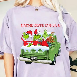 Puede incluir: Camiseta de color lila claro con un diseño festivo. Muestra tres personajes verdes con gorros de Papá Noel en una camioneta verde, con las palabras "DRINK DRANK DRUNK" encima. La camioneta tiene escrito "MERRY CHRISTMAS" y "HO HO HO!"