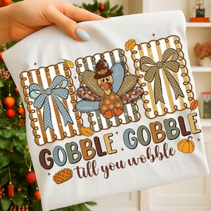 May include: White t-shirt featuring a Thanksgiving-themed design. The design includes a turkey wearing a pilgrim hat, bows, and the text "GOBBLE GOBBLE till you wobble". The design is in shades of brown, blue, and orange.