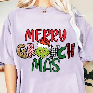 Peut inclure: T-shirt lilas avec l'inscription « MERRY GRINCH MAS » en rouge, vert, noir et blanc. Le personnage du Grinch, portant un bonnet de Père Noël, est au centre. Un design festif pour les fêtes de fin d'année.