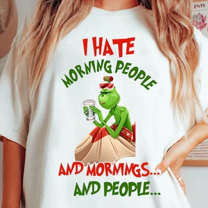 Puede incluir: Camiseta blanca con un gráfico del Grinch sosteniendo una taza de café, con el texto "I HATE MORNING PEOPLE AND MORNINGS... AND PEOPLE..." en rojo y verde. El Grinch viste un atuendo rojo y blanco.