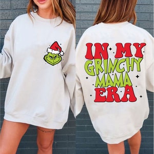 Könnte beinhalten: Cremefarbenes Sweatshirt mit einem Grinch-Motiv mit Weihnachtsmütze auf der Vorderseite. Auf der Rückseite des Sweatshirts steht der Text "IN MY GRINCHY MAMA ERA" in Rot und Grün, mit Sternen.