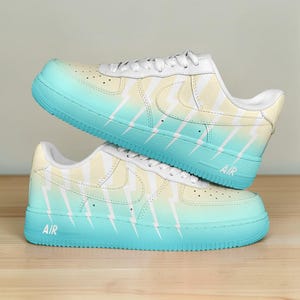 May include: A pair of custom-painted sneakers with a gradient design. The shoes transition from a light yellow to a turquoise blue, featuring white lightning bolt graphics. The word "AIR" is printed on the side of the turquoise sole.