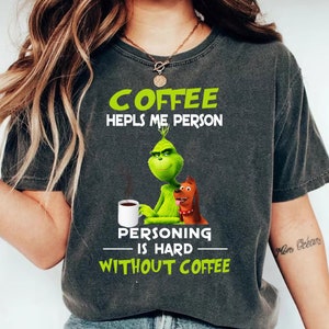 Könnte beinhalten: Dunkelgraues T-Shirt mit einem grünen Grinch-Charakter, der eine Kaffeetasse und einen braunen Hund hält. Das Shirt hat den Text "COFFEE HELPS ME PERSON, PERSONING IS HARD WITHOUT COFFEE" in Grün und Weiß.