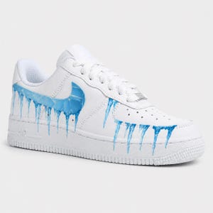 May include: White leather sneaker with a blue ice design. The shoe features a blue Nike swoosh and blue icicles dripping down the side. The sole is white with the word "AIR" in white.