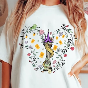 May include: White t-shirt featuring a colorful graphic of a tower with Rapunzel's hair, surrounded by flowers, floating lanterns, and a frog. The design is framed by a floral wreath.