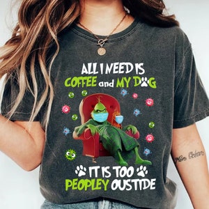 Puede incluir: Camiseta gris oscuro con el texto "ALL I NEED IS COFFEE and MY DOG IT IS TOO PEOPLEY OUSTIDE." El gráfico presenta un personaje verde con mascarilla, sentado en una silla roja y sosteniendo una taza de café. La camiseta tiene un aspecto vintage.