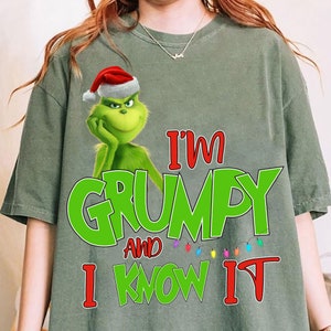 Puede incluir: Camiseta verde salvia con un dibujo animado del Grinch con un gorro de Papá Noel. El texto "I'm Grumpy and I Know It" está impreso en verde y rojo con una cadena de luces de colores. Un diseño divertido y festivo.