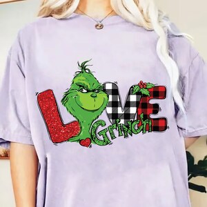Puede incluir: Camiseta lila claro con un gráfico del Grinch y la frase "LOVE Grinch". La letra "L" es roja brillante, la "V" es a cuadros blancos y negros y la "E" es a cuadros rojos.