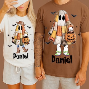 Puede incluir: Camisetas blancas y marrones con un diseño de fantasma de Halloween. El fantasma lleva una capa de patchwork, un sombrero y sostiene una linterna de calabaza. El nombre "Daniel" está impreso debajo. También hay murciélagos y cruces.