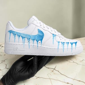 May include: White Air Force 1 sneakers with a custom design featuring blue icicles dripping from the Nike swoosh and along the side. The word "AIR" is visible on the midsole. The shoe is held by a gloved hand.