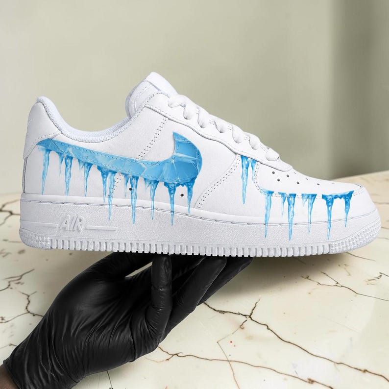 May include: White Air Force 1 sneakers with a custom design featuring blue icicles dripping from the Nike swoosh and along the side. The word "AIR" is visible on the midsole. The shoe is held by a gloved hand.