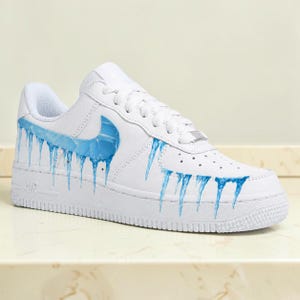 May include: White athletic shoe with a blue swoosh and blue icicle design. The shoe features a white sole, laces, and a perforated toe box. The design is painted on the side and front of the shoe.