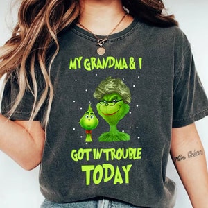 Puede incluir: Camiseta gris oscuro con el texto "MY GRANDMA & I GOT IN TROUBLE TODAY" en verde. La camiseta presenta un personaje de dibujos animados del Grinch con una cara verde, gafas y una peluca, y un personaje del Grinch más pequeño.