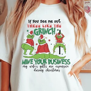 Puede incluir: Camiseta blanca con un gráfico humorístico que presenta a tres personajes de Grinch en varias poses. El texto dice: "If you see me out THREE LIKE THE GRINCH MINE YOUR BUSINESS my wifes gifts are expensive during christmas."