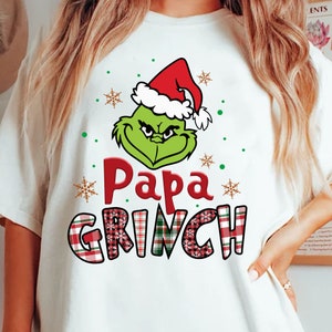Puede incluir: Camiseta blanca con la cara verde del Grinch con un gorro de Papá Noel rojo. Las palabras "Papa Grinch" son rojas, con "Grinch" en un patrón de cuadros. Copos de nieve y puntos completan el diseño navideño.