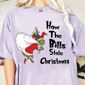 Może przedstawiać: Jasnofioletowa koszulka z kreskówkowym Grinchem niosącym worek z prezentami i choinkę. Tekst "How The Bills Stole Christmas" jest wydrukowany na czarno.