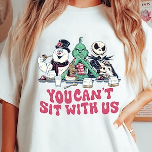 Könnte beinhalten: Weißes T-Shirt mit einer Cartoon-Illustration von Frosty dem Schneemann, dem Grinch und Jack Skellington. Der Text "YOU CAN'T SIT WITH US" ist in Rosa. Die Charaktere sitzen an einem Tisch mit Milch und Snacks.