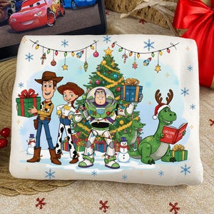 May include: A white rectangular cushion with a festive Toy Story design. Woody, Jessie, Buzz Lightyear, and Rex are shown with presents and a decorated Christmas tree. The cushion is decorated with snowflakes and fairy lights.
