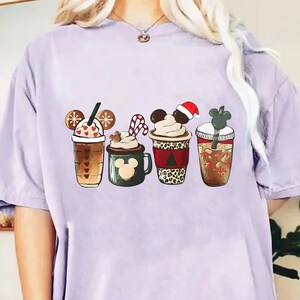 May include: A light purple t-shirt with a graphic of four different holiday-themed coffee cups. The cups are decorated with Mickey Mouse ears, snowflakes, candy canes, and a Santa hat.