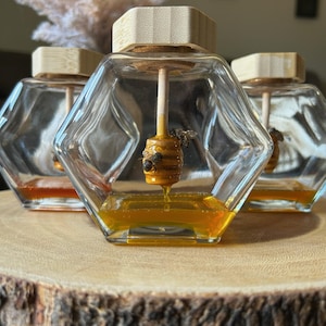 Peut inclure: Trois bocaux en verre hexagonaux avec des couvercles en bois clair, chacun contenant une cuillère à miel en bois suspendue. Une petite cuillère à miel est recouverte de miel doré avec une figurine d'abeille, et du miel liquide au fond.