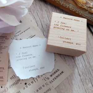 May include: A wooden stamp with the text "< Record Memo >" "I feel the flowers growing on me." | Collect stamps, etc" on it. The stamp is sitting on a piece of paper with the same text.