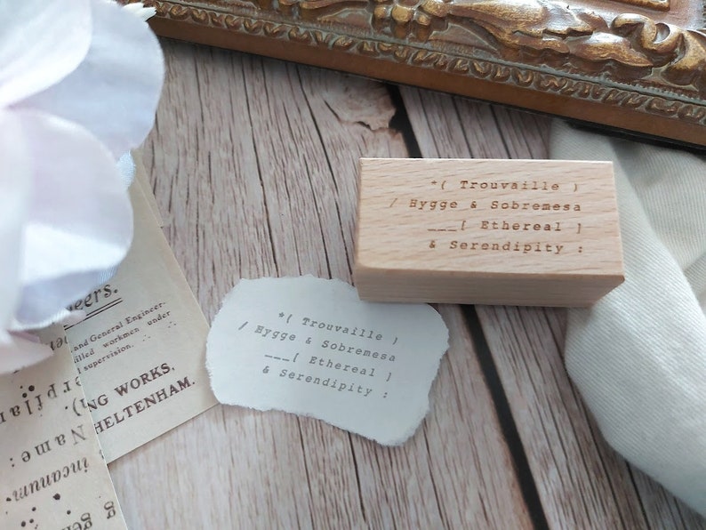 May include: A wooden stamp with the text "*(Trouvaille) / Hygge & Sobremesa --| Ethereal | & Serendipity:"  The stamp is sitting on a wooden surface.