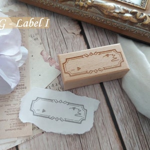 May include: A wooden stamp with a decorative border and a blank space for adding text. The stamp is on a white surface with a piece of paper with a stamped image of the same design.