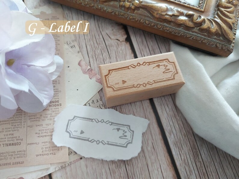 May include: A wooden stamp with a decorative border and a blank space for adding text. The stamp is on a white surface with a piece of paper with a stamped image of the same design.