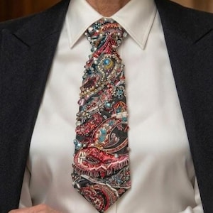 Puede incluir: Primer plano de una corbata estampada con un diseño de cachemira. La corbata está adornada con cuentas y joyas de colores. La corbata se lleva sobre una camisa blanca con cuello y un blazer negro.