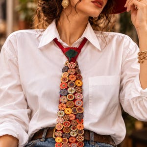 May include: A red necktie adorned with numerous small, round, multi-colored wooden buttons featuring various floral and geometric patterns. The tie is worn over a crisp white button-down shirt, paired with blue denim jeans and a brown leather belt.
