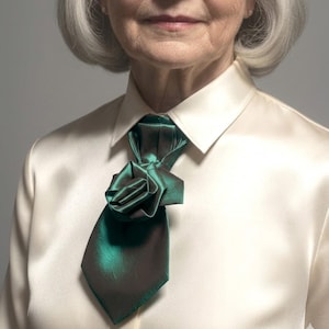 May include: A close-up of a person wearing a cream-colored button-down shirt with a dark green tie. The tie is styled with a rose-like knot at the neck. The tie has a shiny, iridescent finish.
