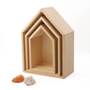 Könnte beinhalten: Ein Satz von vier holzförmigen, hausförmigen Nistregalen in verschiedenen Größen. Die Regale sind aus hellfarbigem Holz gefertigt und so angeordnet, dass sie ineinander passen. Zwei Muscheln befinden sich am Fuß der Regale.