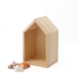 Op de afbeelding: Een klein houten huisvormig rek met een natuurlijke houten afwerking. Het rek is leeg, maar er liggen vier schelpen voor.