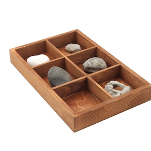 Puede incluir: Una bandeja de madera con seis compartimentos, cada uno con una piedra diferente. Las piedras varían en forma, tamaño y color, incluyendo variedades blancas, grises y moteadas. La bandeja es de madera marrón claro.