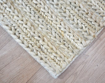 May include: Close-up of a natural fiber rug with a woven, braided design. The rug is a light beige color and has a textured surface. The corner of the rug is visible, showing the edge and the weave pattern. The rug is on a wooden floor.