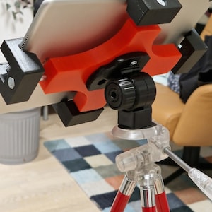 May include: A red and black tablet stand with a suction cup base attached to a red tripod. The stand is designed to hold a tablet securely in place.