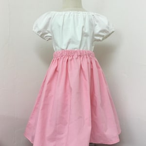 Può includere: Abito a due pezzi per bambina. La parte superiore è bianca con maniche corte a sbuffo e scollatura a spalle scoperte. La gonna è rosa acceso, arricciata in vita e svasata. L'abito è esposto su un manichino.
