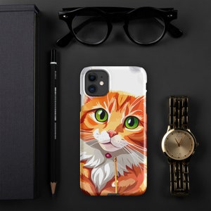 May include: A white iPhone case with a cartoon illustration of a ginger cat with green eyes. The cat is looking up and has a pink jewel around its neck.