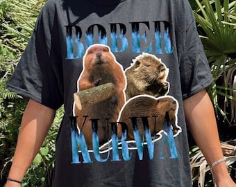 May include: Black t-shirt featuring a beaver graphic with the text "BOBER KURWA" in blue and white. The graphic includes three beaver illustrations. The shirt is worn outdoors with green foliage in the background.