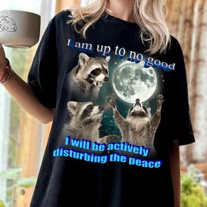 May include: Black t-shirt featuring a graphic of three raccoons under a full moon. The text "I am up to no good" and "I will be actively disturbing the peace" are printed in blue. The shirt is a casual, oversized style.