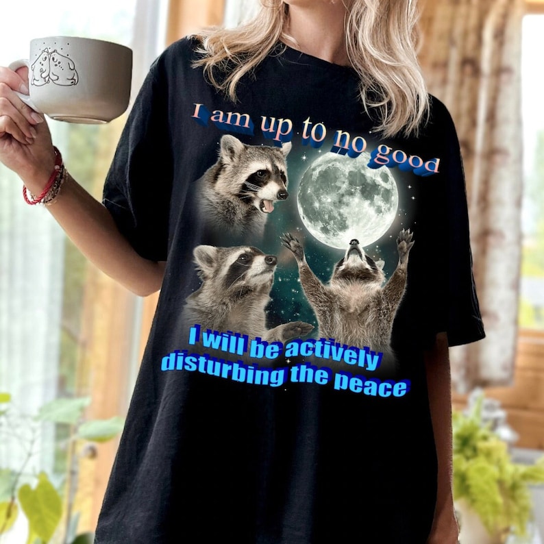 May include: Black t-shirt featuring a graphic of three raccoons under a full moon. The text "I am up to no good" and "I will be actively disturbing the peace" are printed in blue. The shirt is a casual, oversized style.