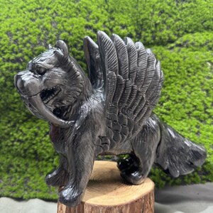 Op de afbeelding: Een donkergrijze sculptuur van een gevleugelde sabeltandkat. Het beeldje heeft gedetailleerde vleugels, een lange staart en prominente hoektanden. Het wordt getoond op een kleine, ronde houten basis, tegen een groene, getextureerde achtergrond.