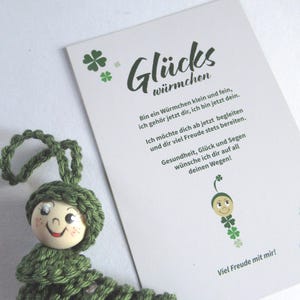 Könnte beinhalten: Ein handgefertigtes grünes gehäkeltes Raupenspielzeug mit einem Holzkopf und einem lächelnden Gesicht. Eine weiße Karte mit deutschem Text und einer vierblättrigen Kleeblattgrafik befindet sich im Hintergrund. Die Karte wünscht Gesundheit, Glück und Segen.