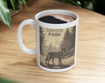 May include: White ceramic mug with a handle, filled with dark coffee. The mug features a graphic of a coyote in a park setting with the text "COYOTE PARK" in a vintage style. The mug is on a wooden surface.