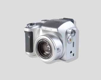 Pode incluir: Uma câmara digital FinePix prateada com uma lente proeminente com zoom ótico de 6x. O corpo da câmara inclui uma pega preta no lado esquerdo, um botão de disparo no topo e várias portas no painel lateral para conetividade e energia.