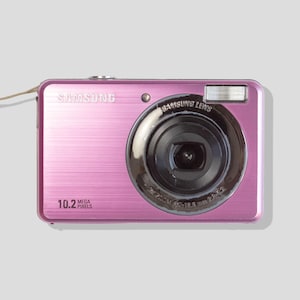 May include: A metallic pink Samsung digital camera featuring a 10.2 megapixels label on the front, a black lens housing with Samsung Lens text, a built-in flash, and a light-colored wrist strap attached to the side of the compact rectangular body.